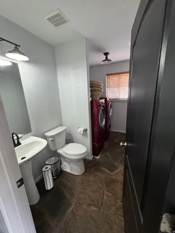 Pedestal sink with a black-finish faucet, toilet, red-finish front-load washer and dryer, and wood-finish blinds