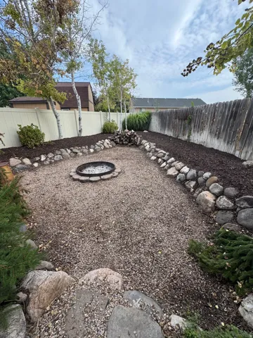 Expansive backyard featuring a gravel patio, built-in fire pit, perimeter landscaping with natural rock accents, and mature trees