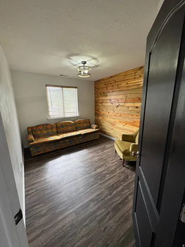 Wood-finish flooring, accent wall featuring horizontal wood planks, window with blinds, ceiling-mounted light fixture, and a dark paneled door