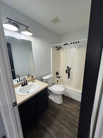 Modern full bathroom featuring dual vanity with wood-finish countertop and round vessel sinks, matte black hardware and mirror fixtures, combined shower-tub with glass enclosure and neutral tile surround, wood-finish flooring throughout, and recessed ceil