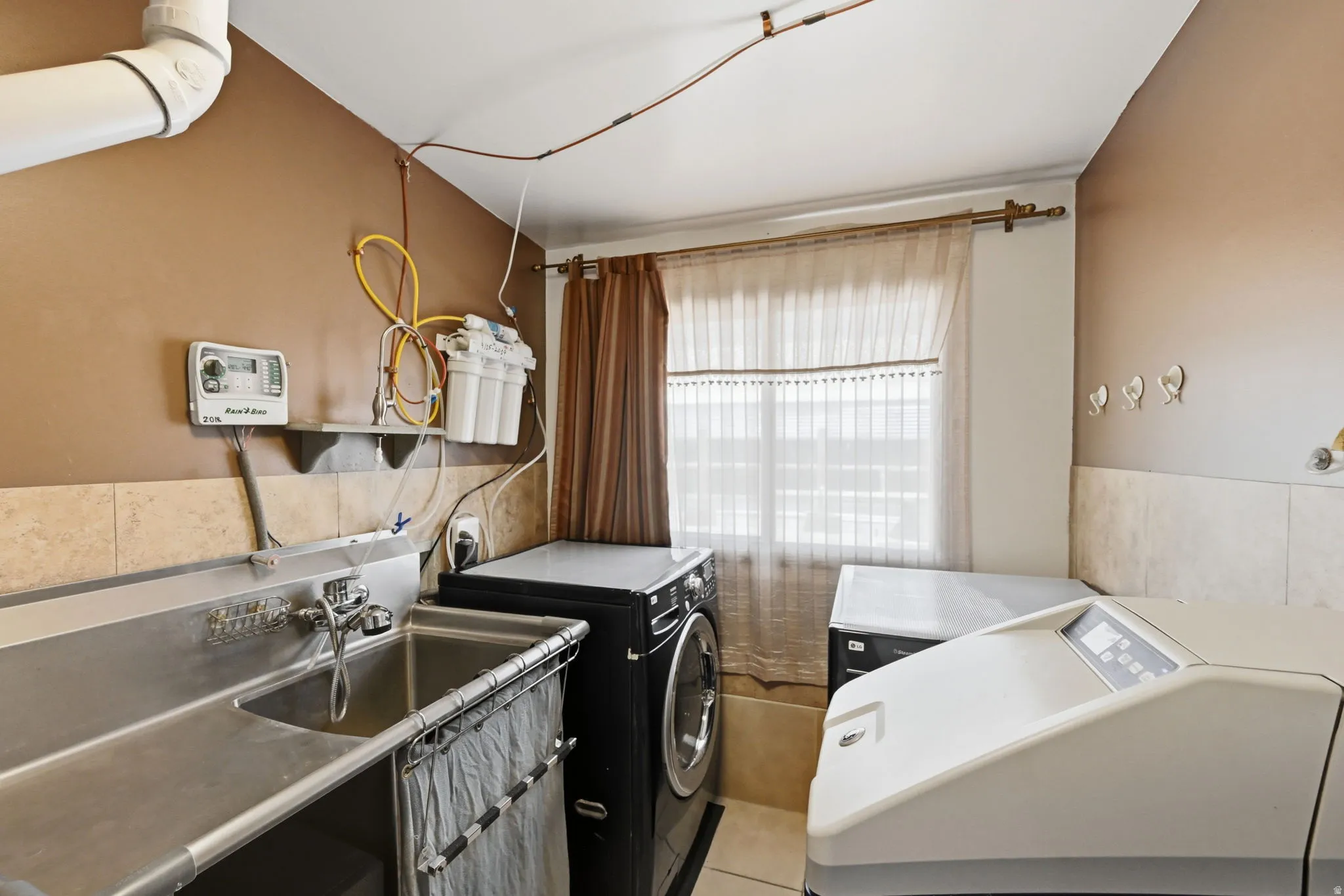 Utility room with a stainless steel sink, tiled backsplash, and window with privacy coverings