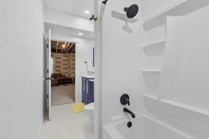White tiled flooring, recessed lighting, matte black shower fixtures, blue vanity with gold hardware, and a white integrated tub