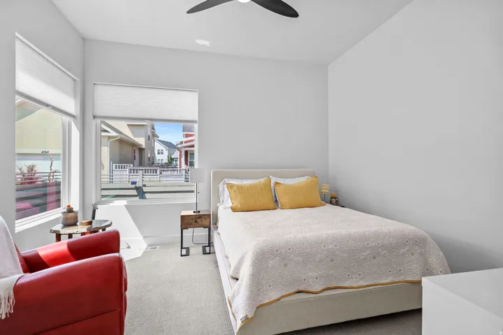 Bright bedroom featuring light-toned walls, two large windows with blinds, a ceiling fan, and carpeted flooring