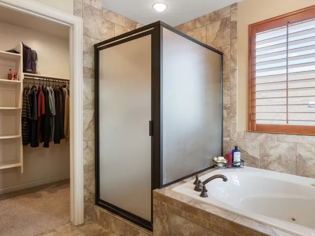 Primary en-suite with Separate built-in jetted tub with tile surround, enclosed shower with frosted glass, wood-framed window with plantation shutters, and an adjacent walk-in closet with shelving and hanging rods.