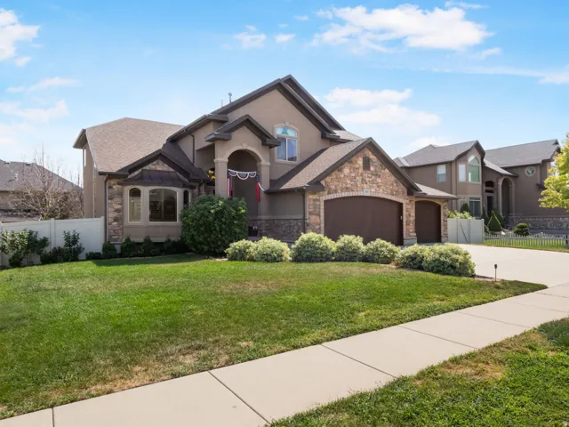 Stucco exterior with stone accents, three-car garage, manicured lawn, paved driveway, and concrete sidewalk.