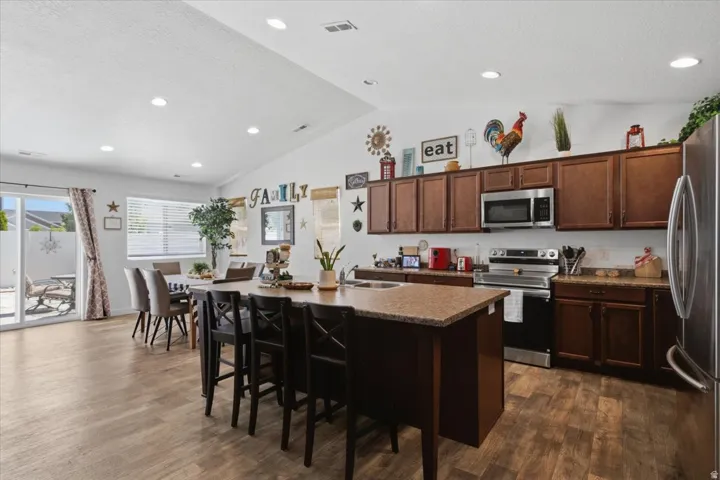 Open-concept kitchen featuring wood-finish flooring, dark wood cabinetry, stainless steel appliances, and a central island with an integrated sink