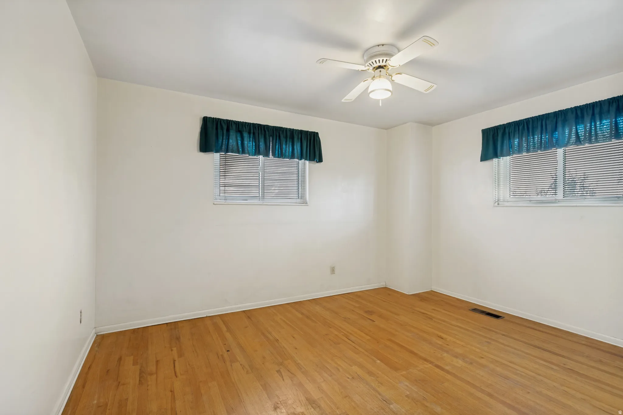 Light-toned wood flooring, bright white walls, two windows with horizontal blinds, and a ceiling fan with integrated lighting
