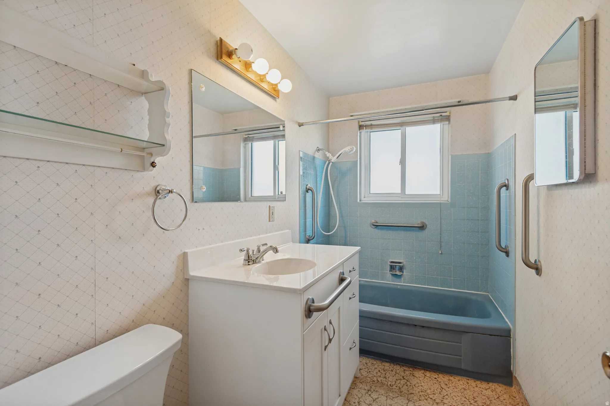 Bathroom featuring a white vanity with integrated sink, a rectangular mirror, a light fixture with four bulbs, a window, and a bathtub with blue wall tile