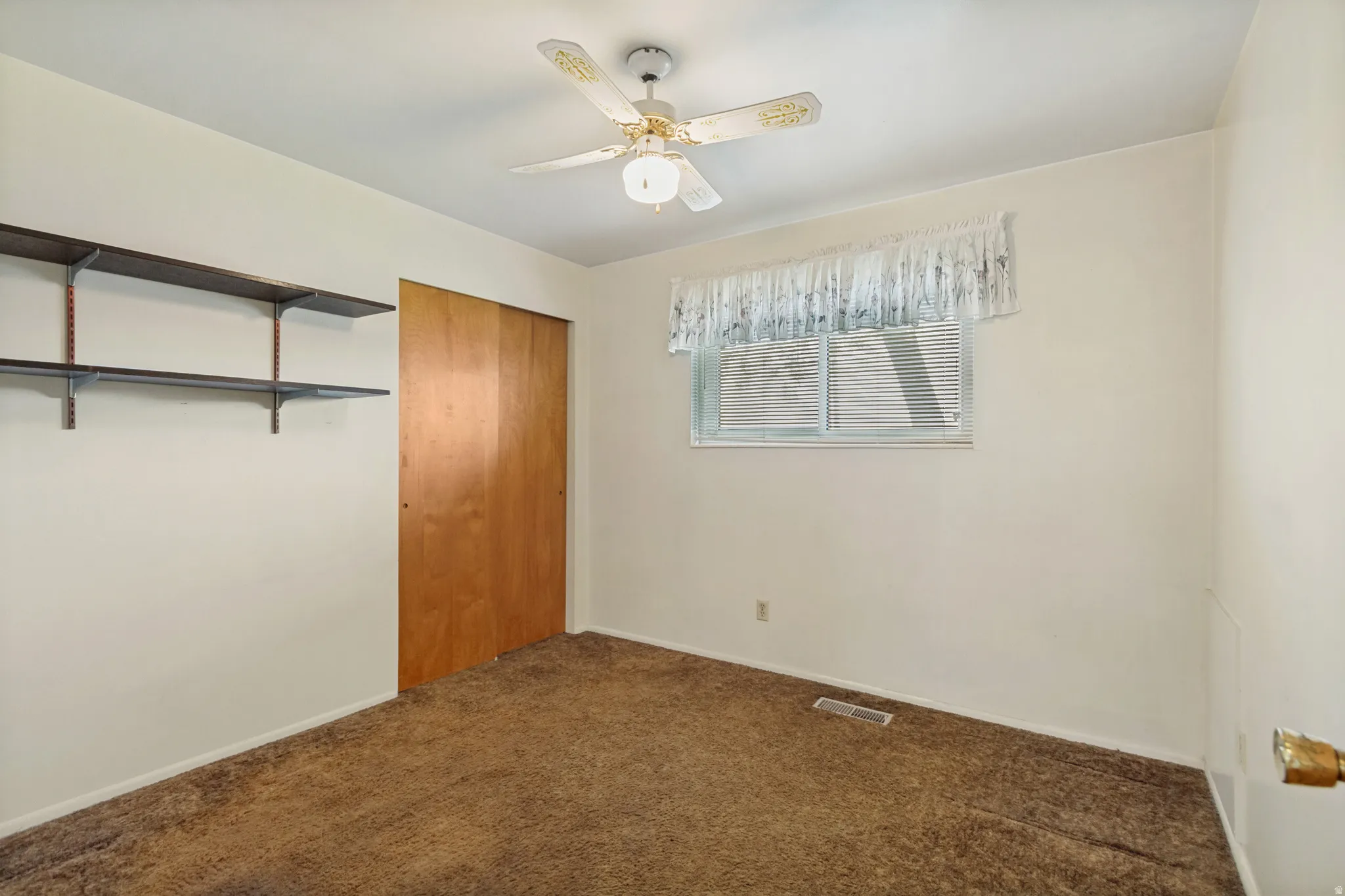 Ceiling fan with light fixture, sliding wood-panel closet doors, window with blinds, and wall-mounted shelving