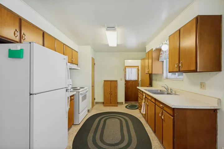 Galley kitchen featuring wood-finish cabinetry, white appliances, a double basin sink, and laminate countertops