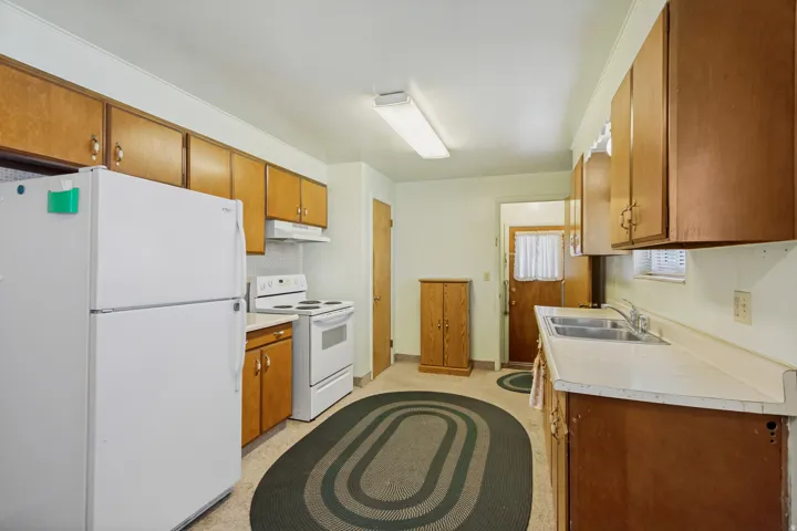 Galley kitchen featuring wood-finish cabinetry, white appliances, dual basin stainless steel sink, and light-toned countertops