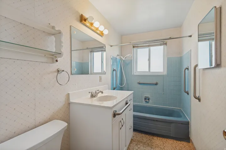 Bathroom featuring a white vanity with integrated sink, a rectangular mirror, a light fixture with four bulbs, a window, and a bathtub with blue wall tile