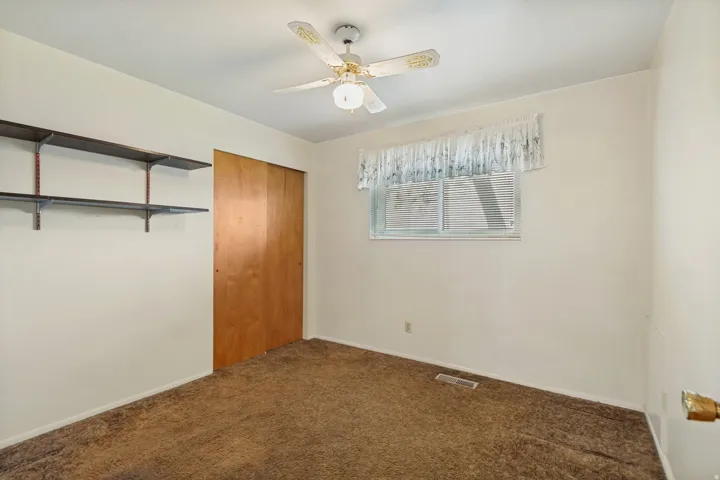 Ceiling fan with light fixture, sliding wood-panel closet doors, window with blinds, and wall-mounted shelving
