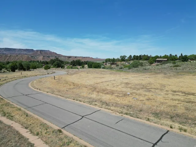 Paved road with curb, expansive undeveloped lot, distant mountain backdrop, scattered trees, blue sky