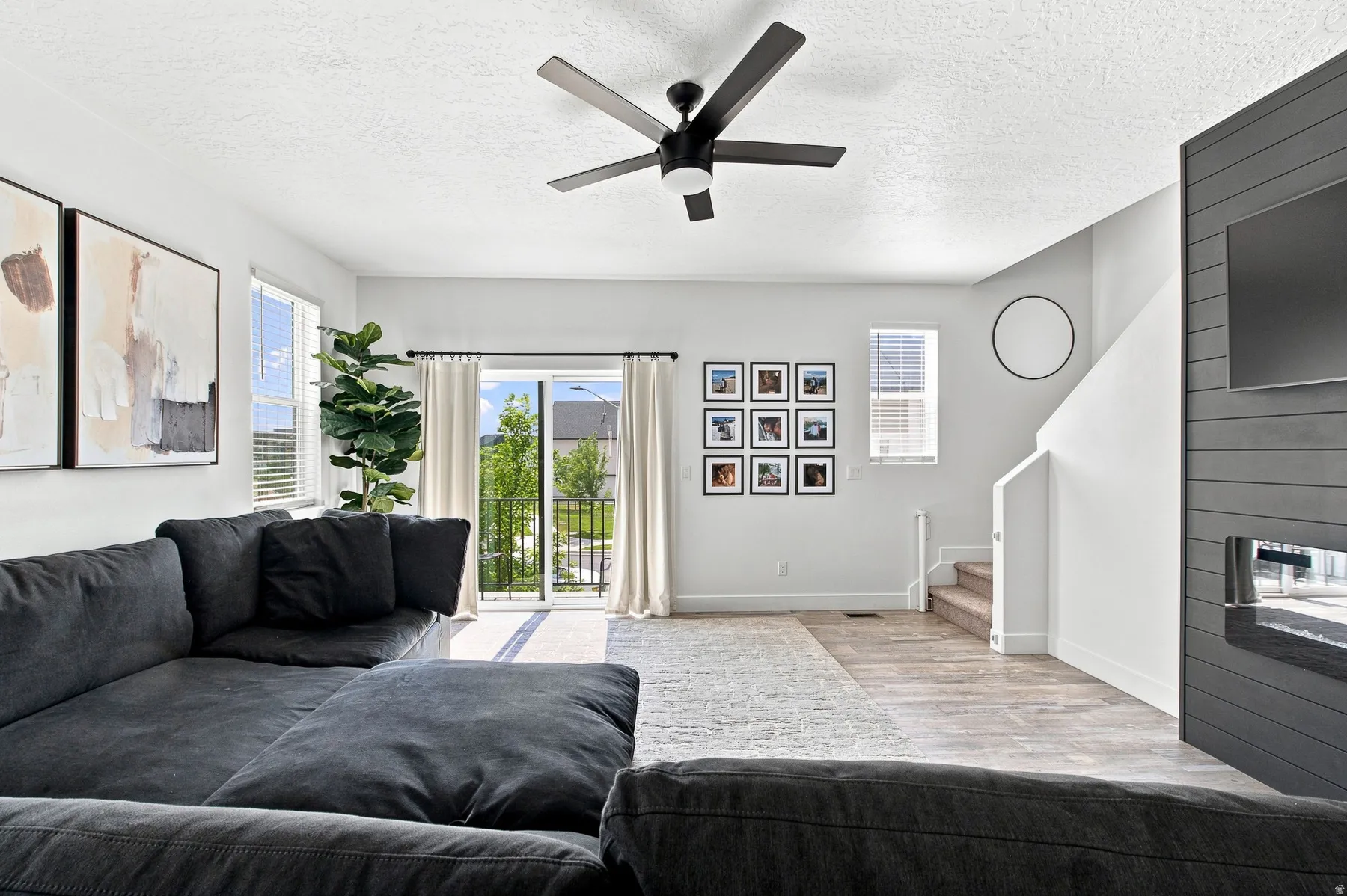 Open-concept living area with wood-finish flooring, a modern black ceiling fan with integrated lighting, and a contemporary fireplace with a dark shiplap surround