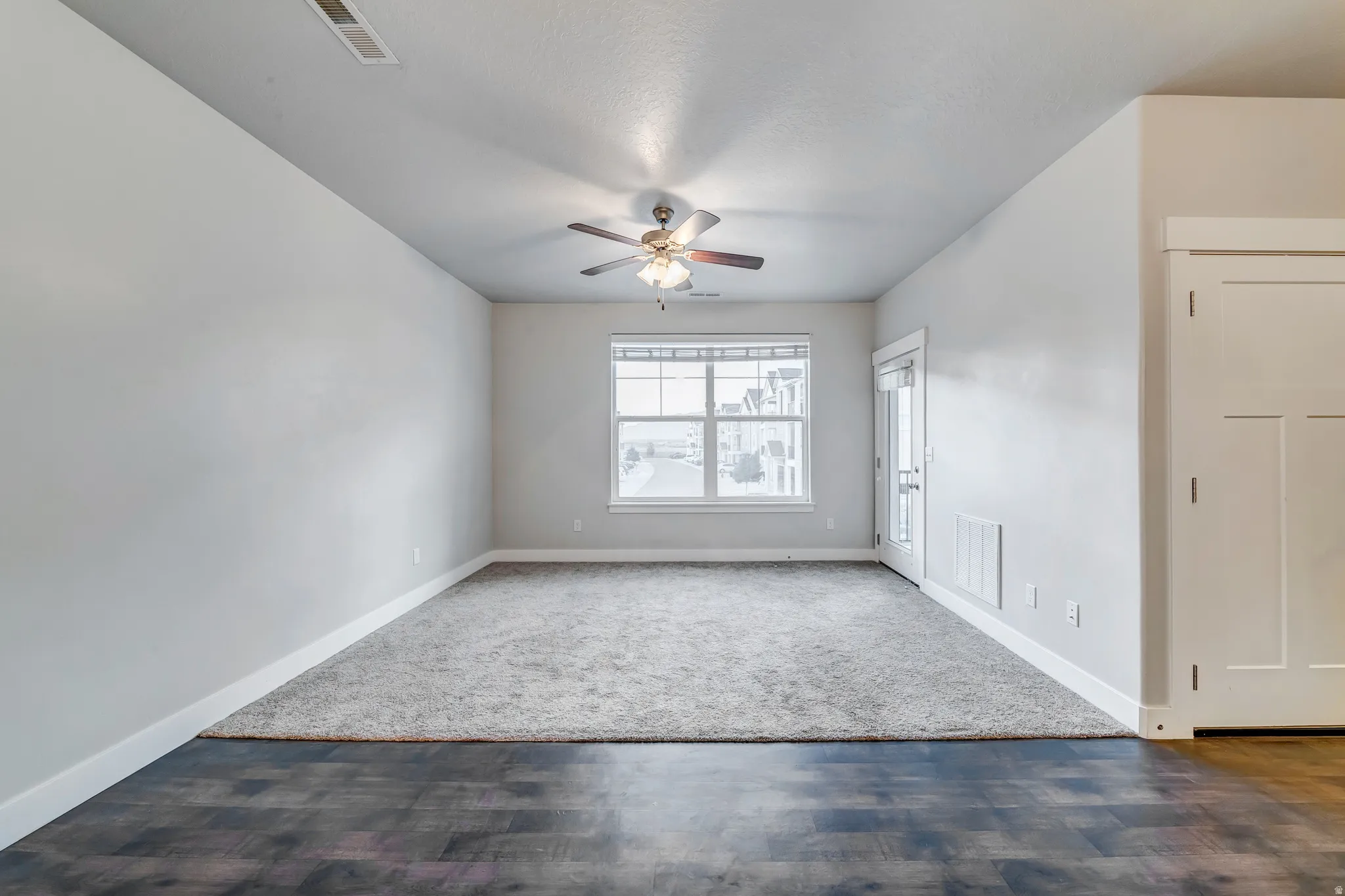 Spacious bedroom with ceiling fan, large double-hung window providing natural light, hardwood and carpet flooring, and generous wall space ideal for furniture arrangement