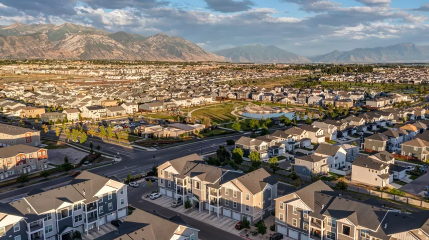 Aerial perspective showcasing a residential community with varied architectural styles, a central park with a pond, and distant mountain ranges