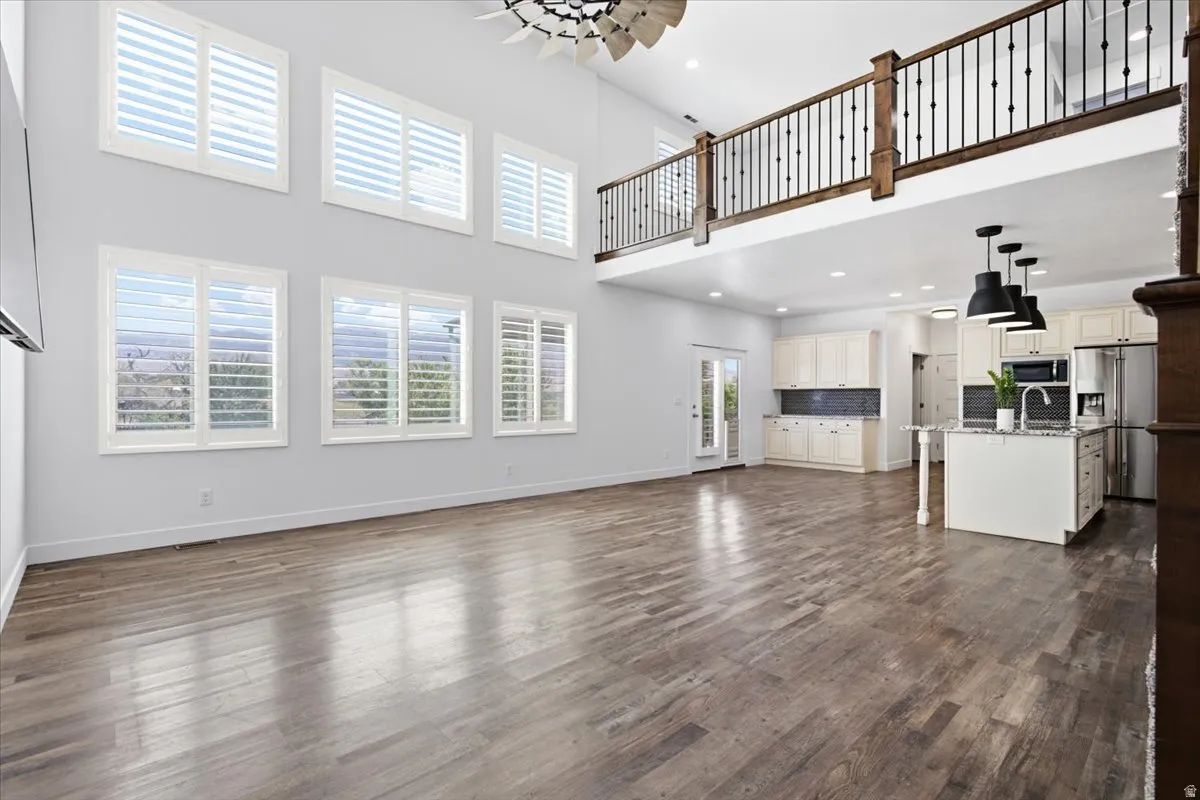 Expansive open-concept living space with wood-finish flooring, two-story windows with plantation shutters, a second-story balcony with wood and wrought iron railing, and a kitchen featuring white cabinetry and a center island with pendant lighting