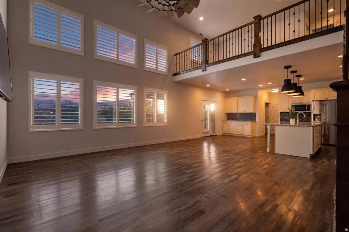 Open-concept living space featuring wood-finish flooring, a kitchen island with pendant lighting, white cabinetry, a stainless steel refrigerator, and a loft with a wrought iron railing