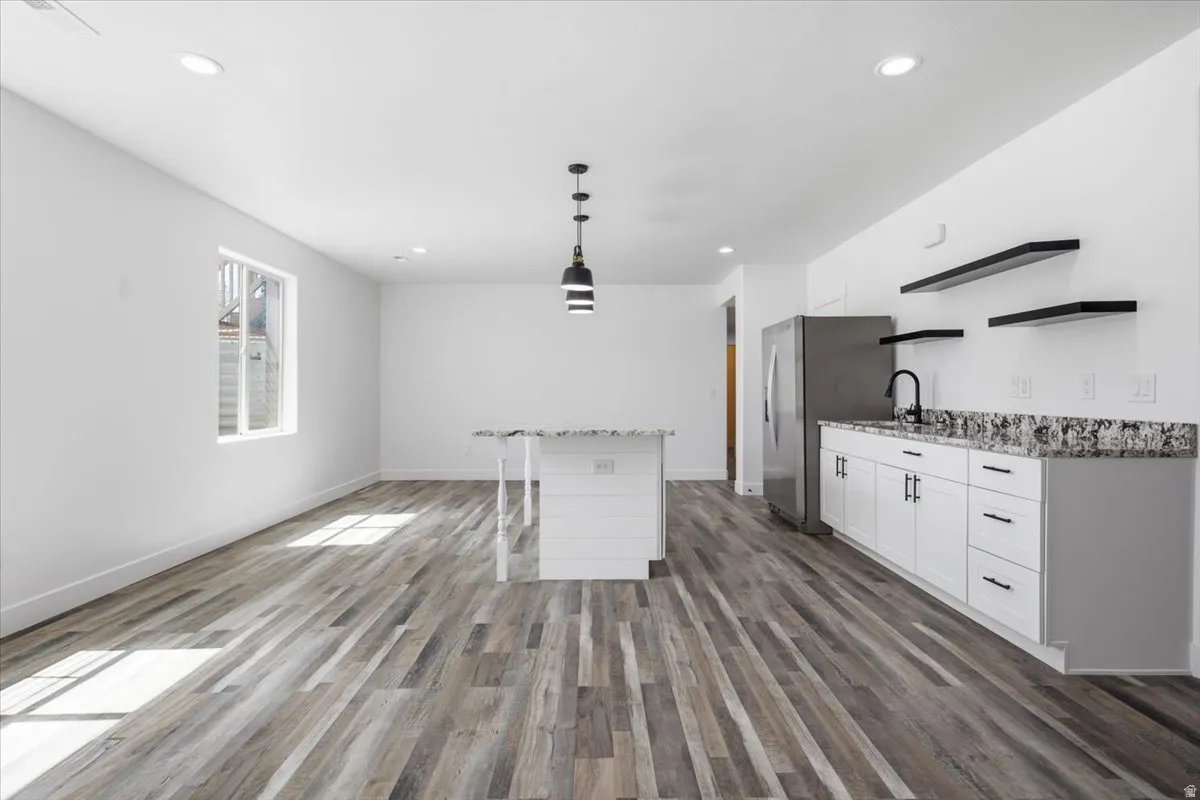 Open-concept living space featuring wood-finish flooring, recessed lighting, a kitchen island with a stone-finish countertop, white cabinetry with dark hardware, and a stainless steel refrigerator