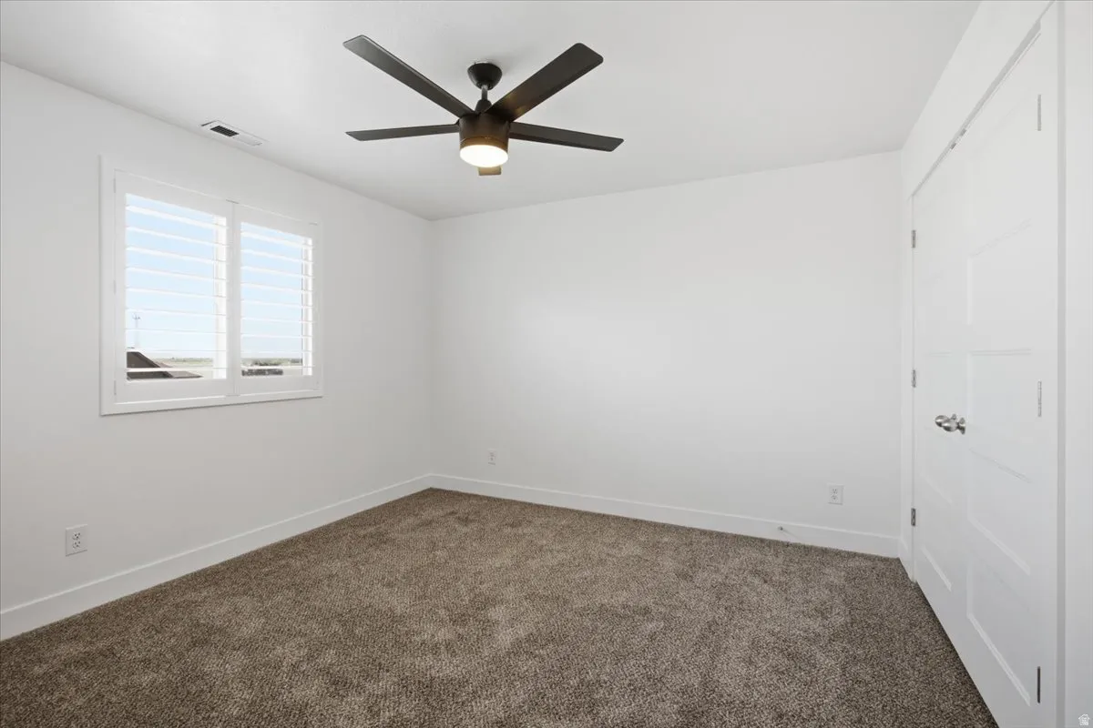 White interior walls, plush carpeting, ceiling fan with integrated lighting, window with white plantation shutters, and a paneled interior door