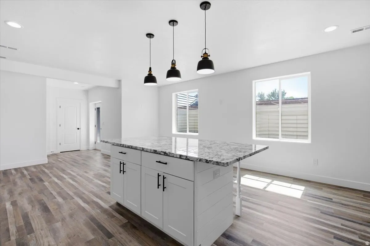 Central island with a stone countertop, three pendant light fixtures, wood-finish flooring, and white walls