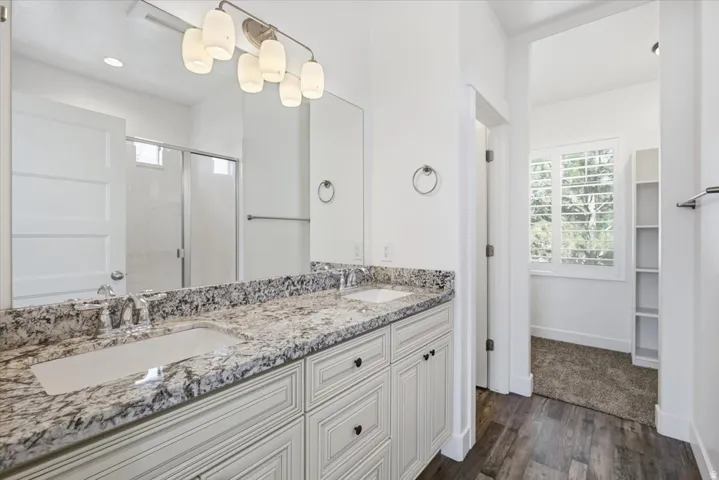 Dual vanity with stone-finish countertop, wood-finish flooring, built-in shelving, and a shower enclosure