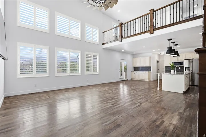 Expansive open-concept living space with wood-finish flooring, two-story windows with plantation shutters, a second-story balcony with wood and wrought iron railing, and a kitchen featuring white cabinetry and a center island with pendant lighting