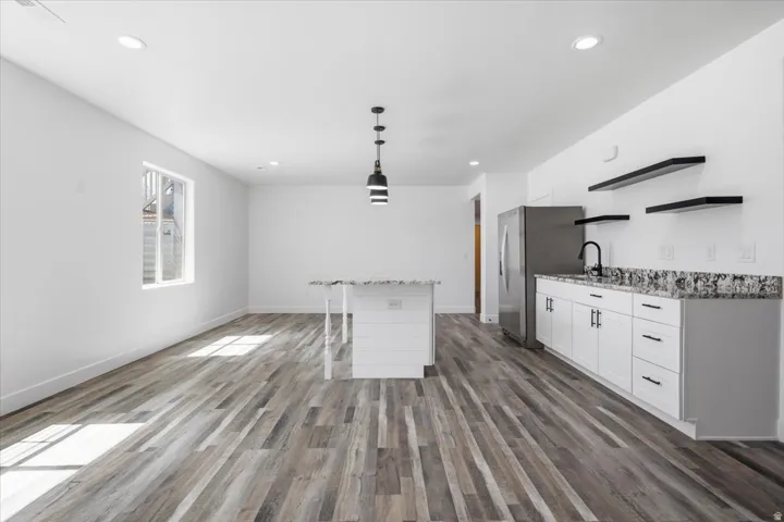 Open-concept living space featuring wood-finish flooring, recessed lighting, a kitchen island with a stone-finish countertop, white cabinetry with dark hardware, and a stainless steel refrigerator