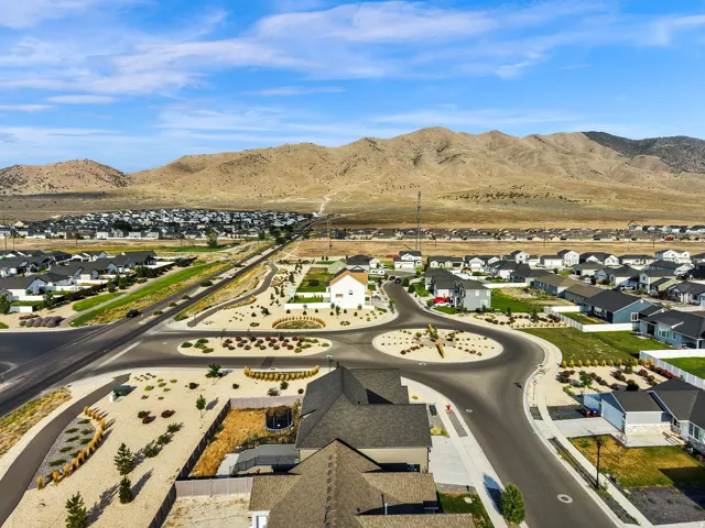 Aerial view of a newly developed residential community featuring modern single-family homes with contemporary architecture, finished landscaping including desert plantings and mature trees, paved streets with curved design elements, and mountain backdrop