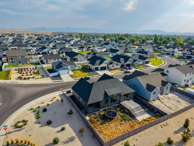Aerial view of a modern residential estate featuring a striking contemporary home with a dark pitched roof, expansive driveway with decorative hardscaping, and manicured landscaping with ornamental plantings