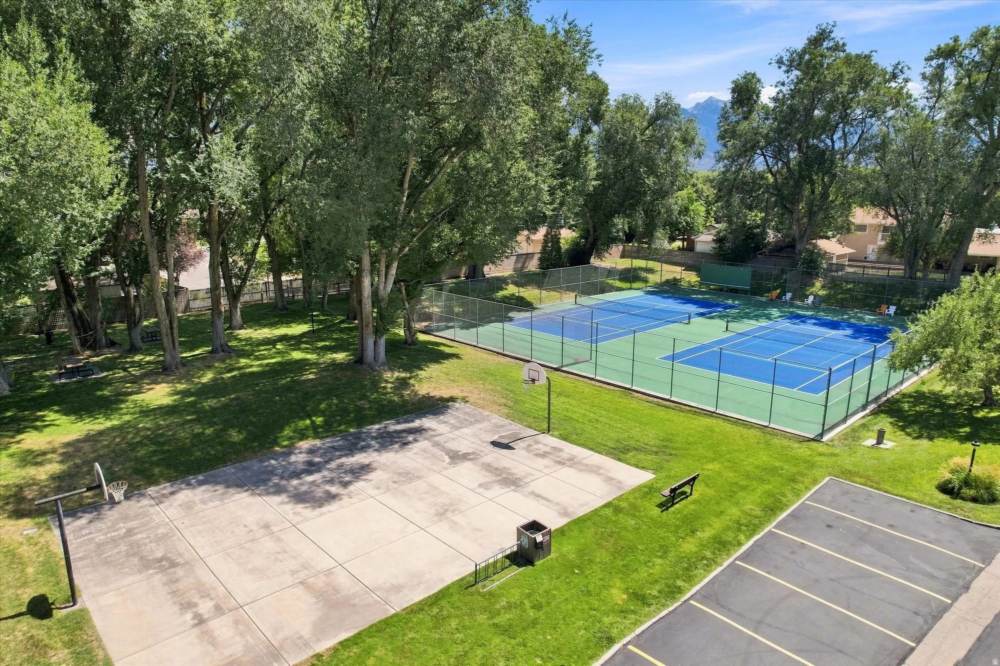 Expansive outdoor amenities featuring two fenced tennis courts, a basketball court, and a paved parking area, all surrounded by mature trees and green spaces