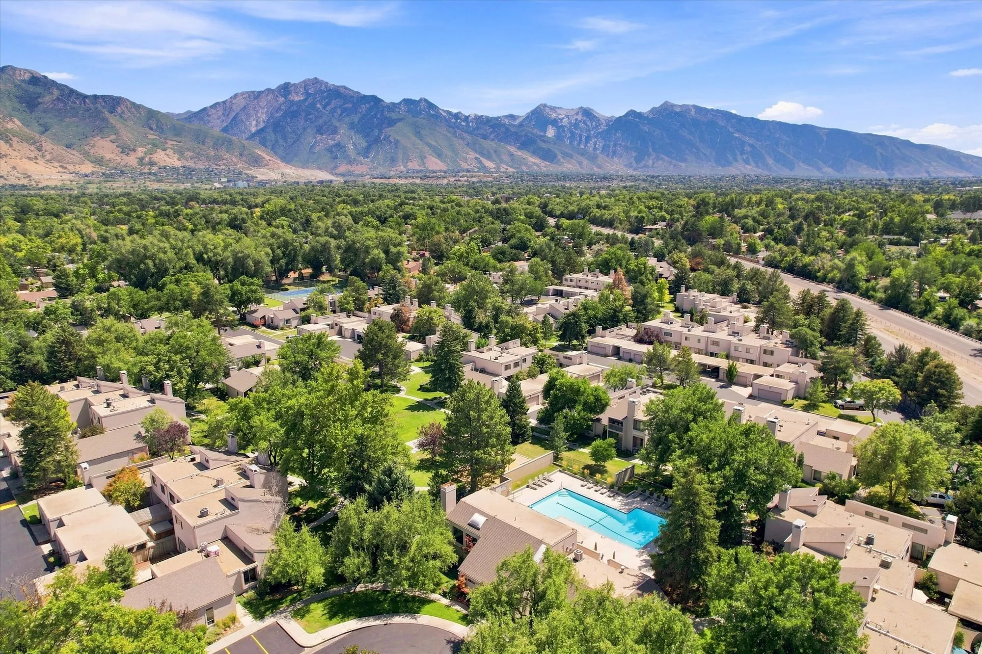 Community buildings with sloped roofs, surrounded by dense mature trees, featuring an outdoor swimming pool, and framed by distant mountain ranges