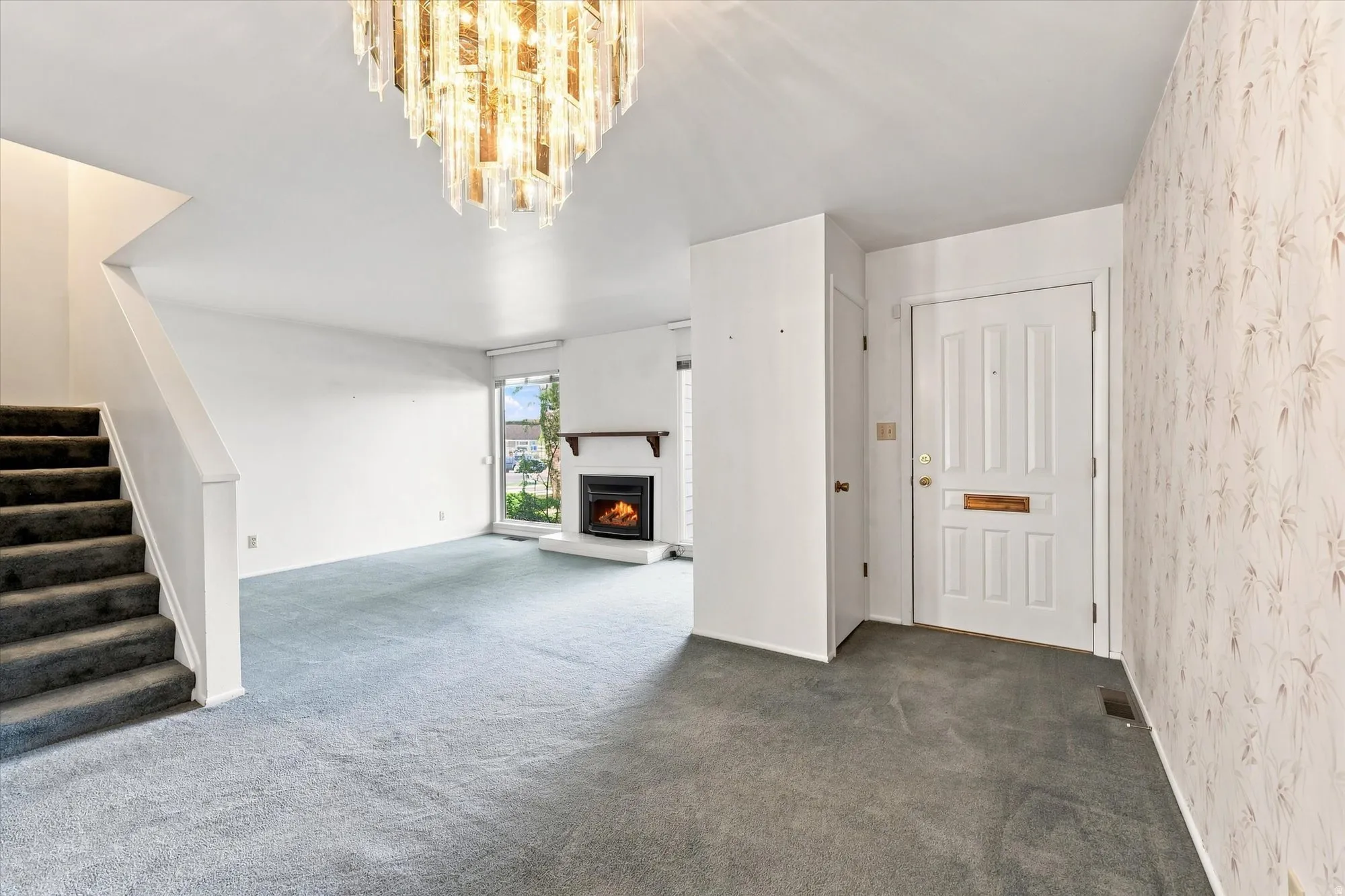 Carpeted living space featuring a fireplace, a large window, a staircase, and a decorative chandelier