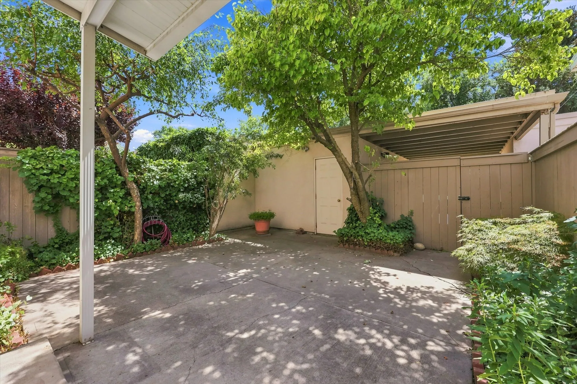 Paved outdoor area featuring a partial overhead covering, mature trees, wood fencing, and a built-in storage shed