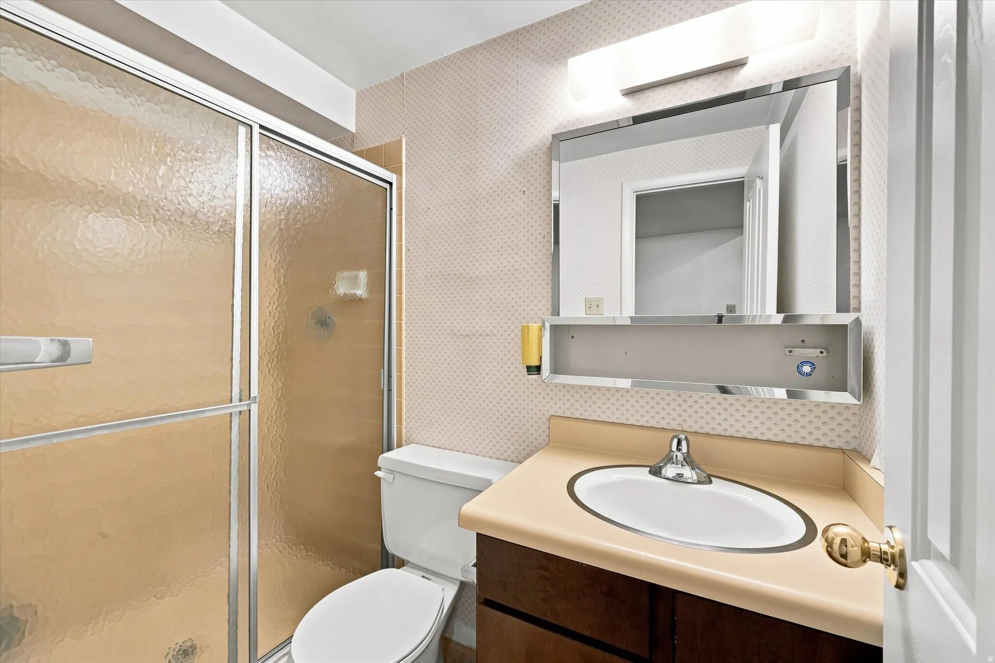 Frosted glass shower enclosure, chrome-framed mirror with integrated lighting, wood-finish vanity, and a neutral countertop with an integrated sink