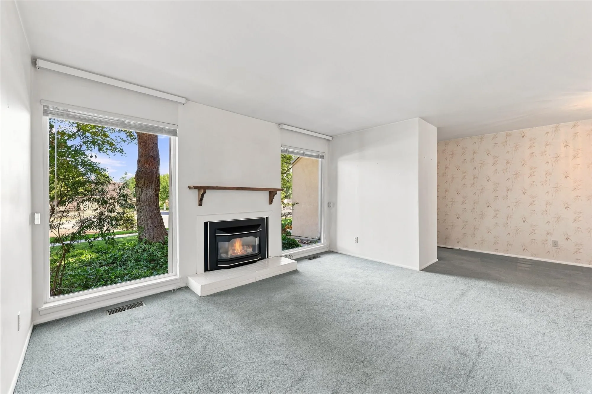 Recessed fireplace with a white hearth, large windows, light-colored carpeting, and wallpapered accent wall