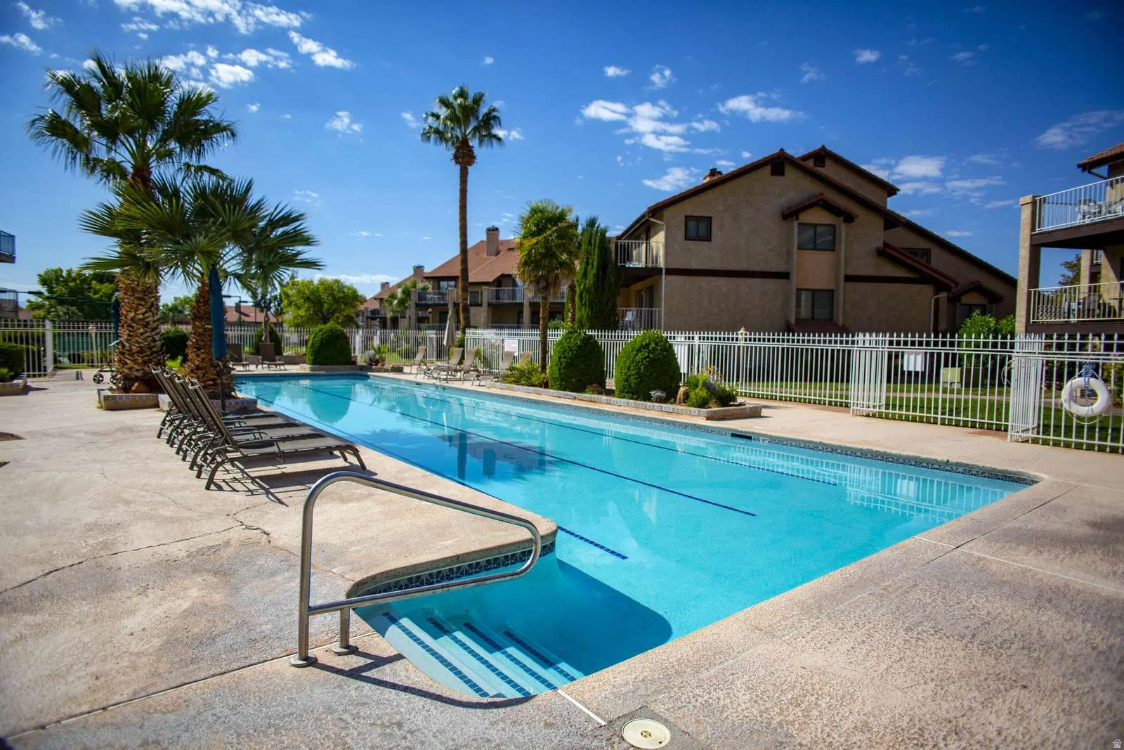 Community swimming pool with concrete decking, surrounded by palm trees and shrubbery, featuring white perimeter fencing