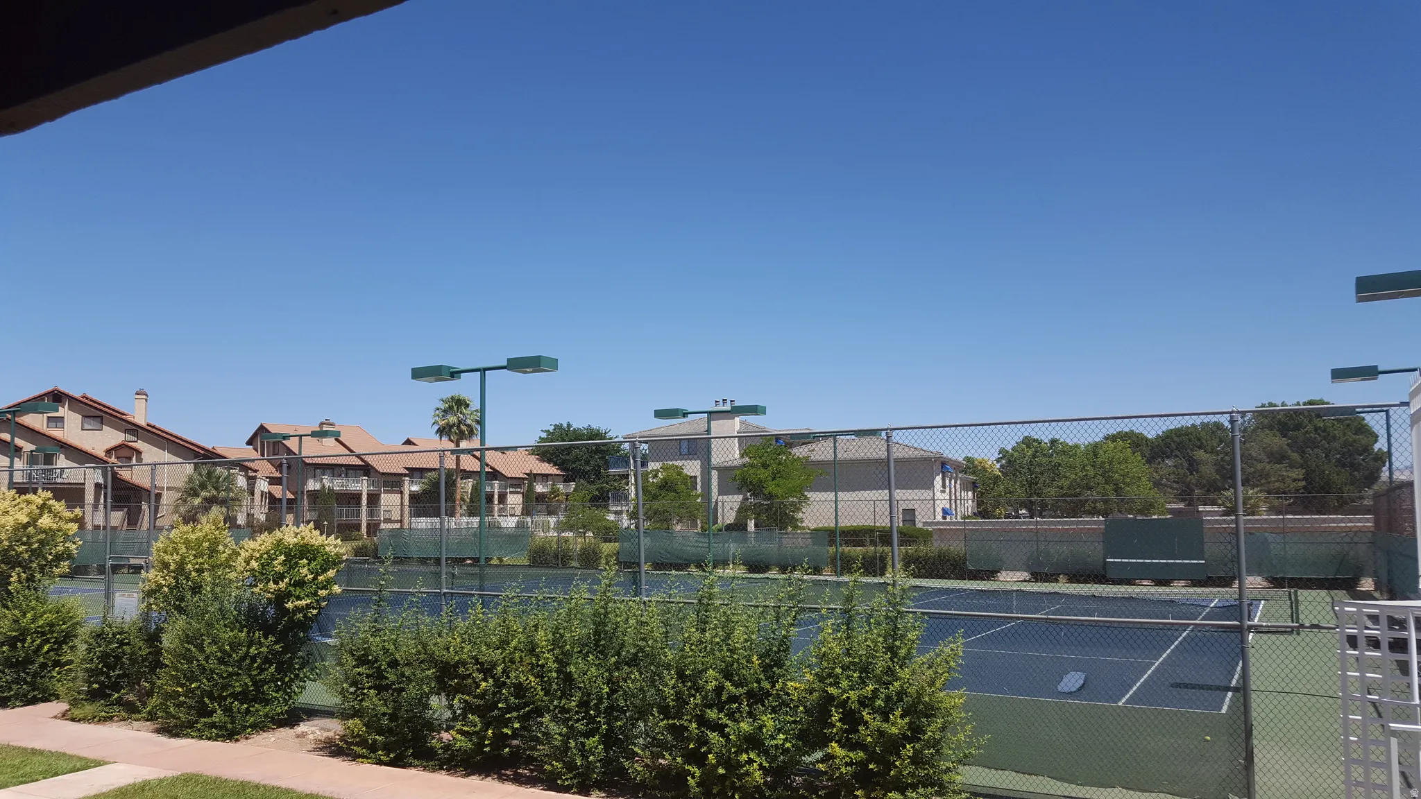 Full-size tennis court with a chain-link fence, surrounded by mature landscaping and residential buildings