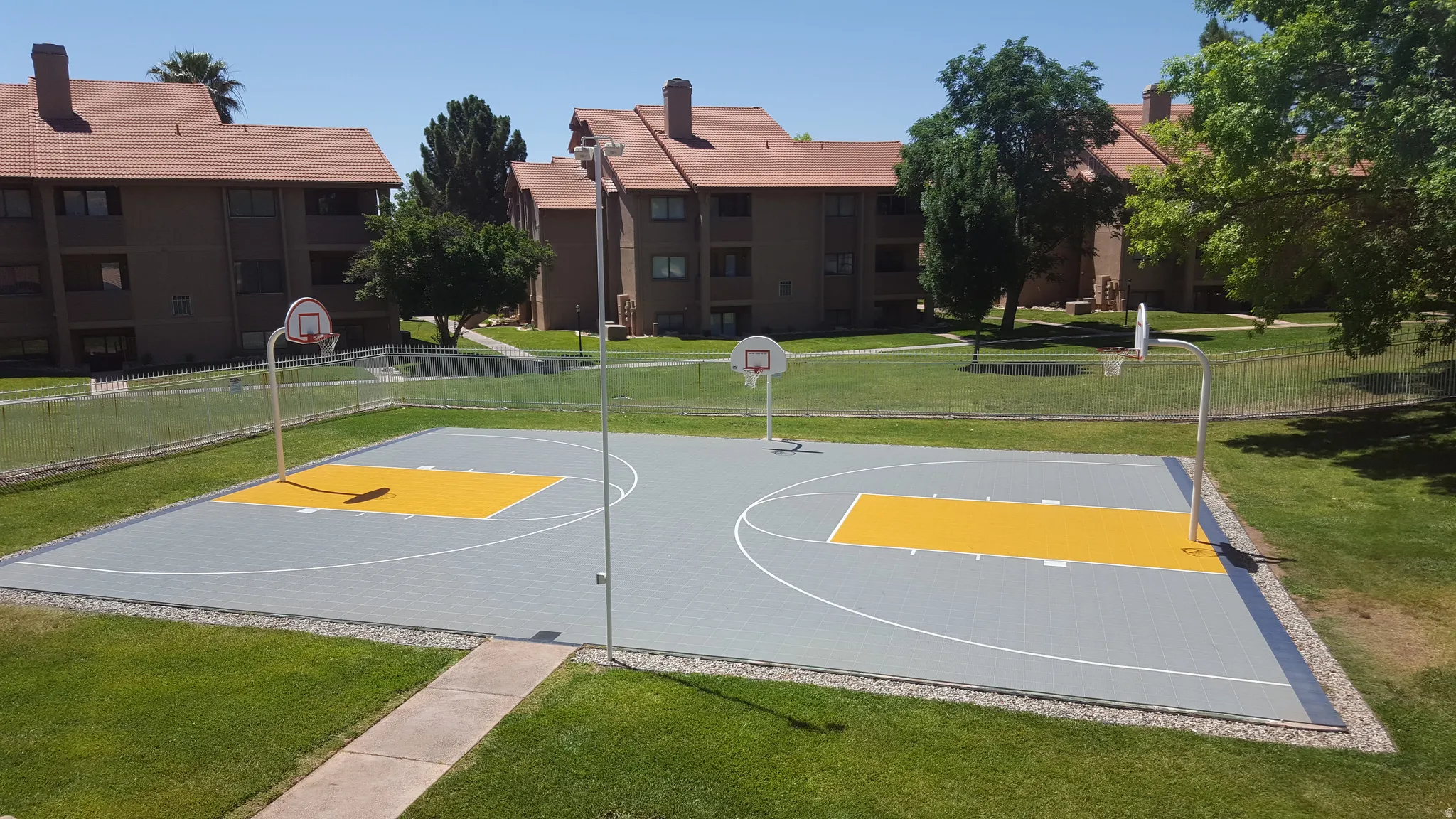 Recreational basketball court with two hoops, surrounded by a chain-link fence and green lawn