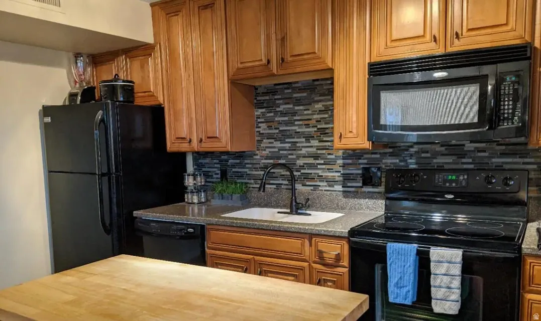 Kitchen featuring wood cabinetry, black appliances, a textured tile backsplash, a single-basin sink with a matte black faucet, and a light wood-finish countertop