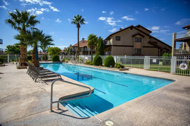Community swimming pool with concrete decking, surrounded by palm trees and shrubbery, featuring white perimeter fencing