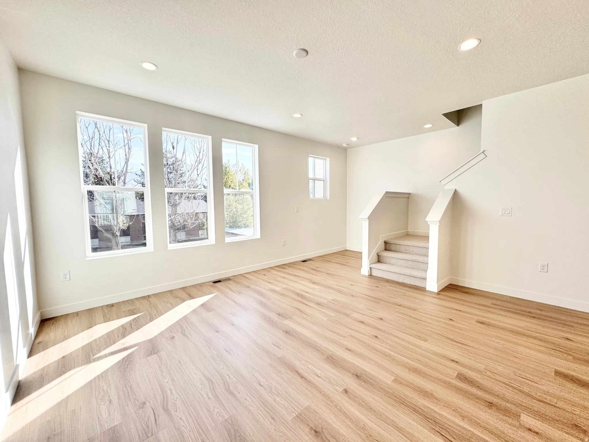 Spacious interior with wood-finish flooring, recessed lighting, multiple windows, and a carpeted staircase with white railings