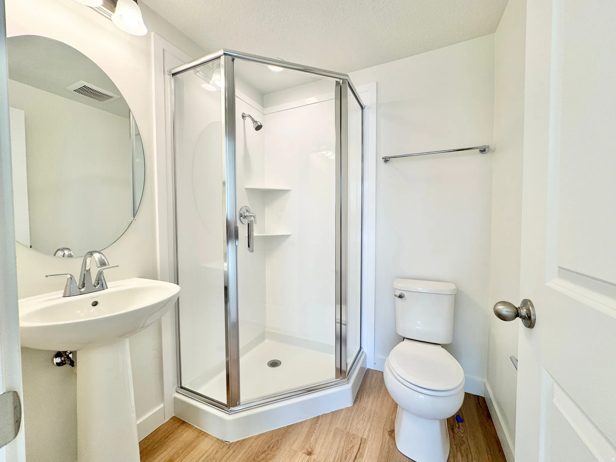 Pedestal sink with chrome faucet, glass-enclosed shower, white toilet, wood-finish flooring, and round wall mirror