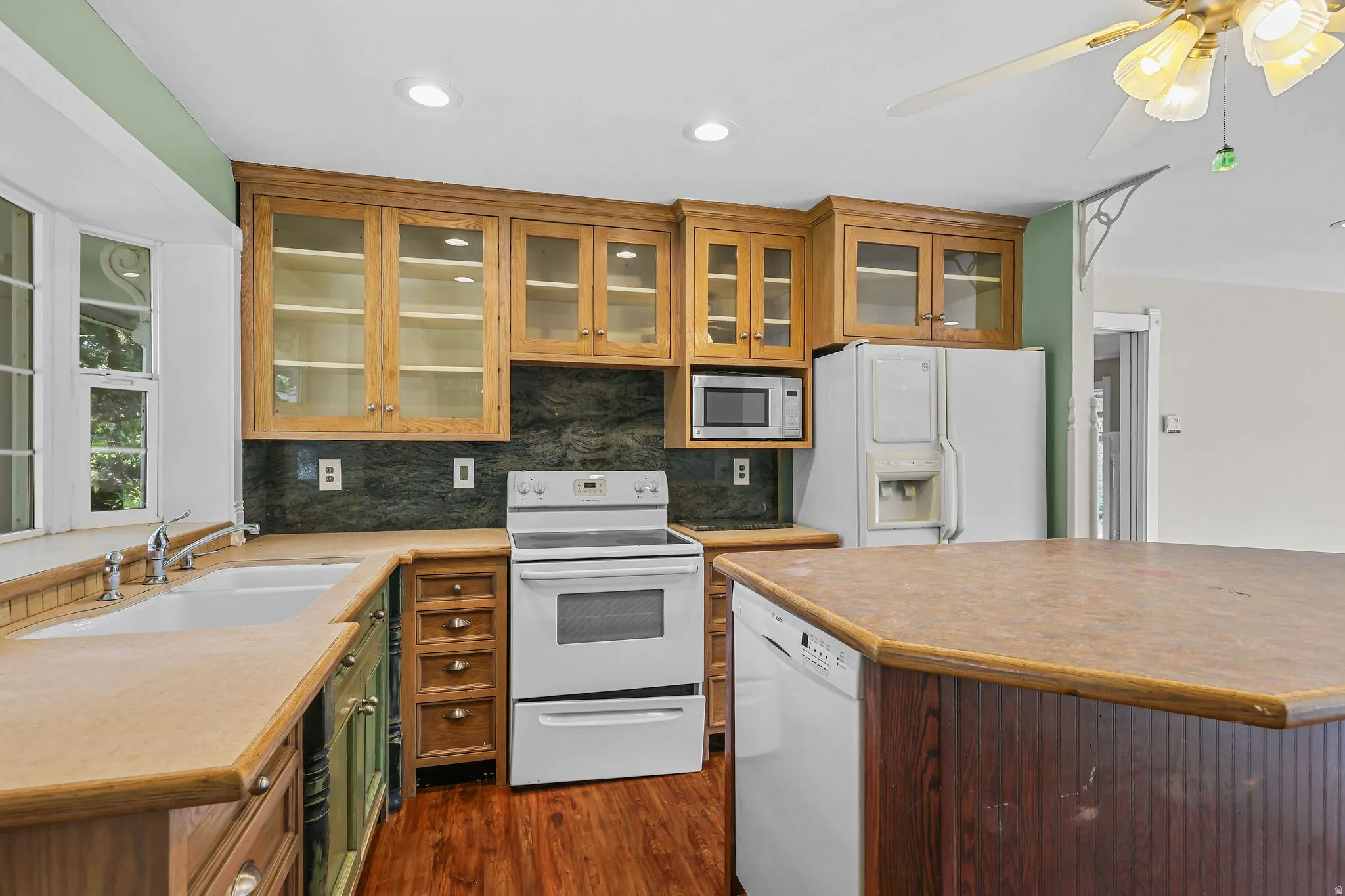 Wood-finish flooring, wood cabinetry with glass-front uppers, dual basin sink with gooseneck faucet, built-in microwave, and ceiling fan with integrated lighting