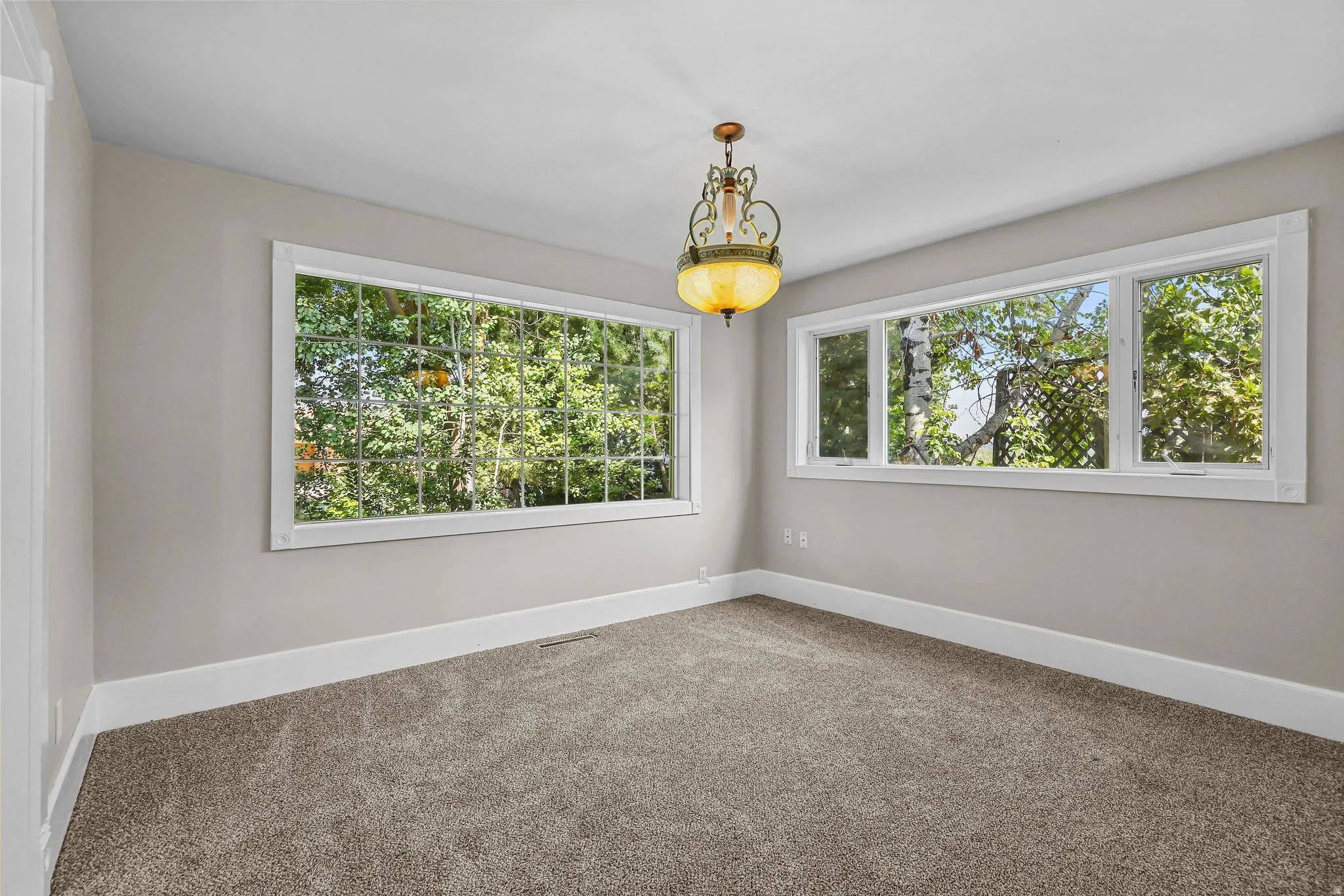 Bright corner room with expansive windows framing mature tree-lined scenery, neutral carpet flooring, and crisp white trim