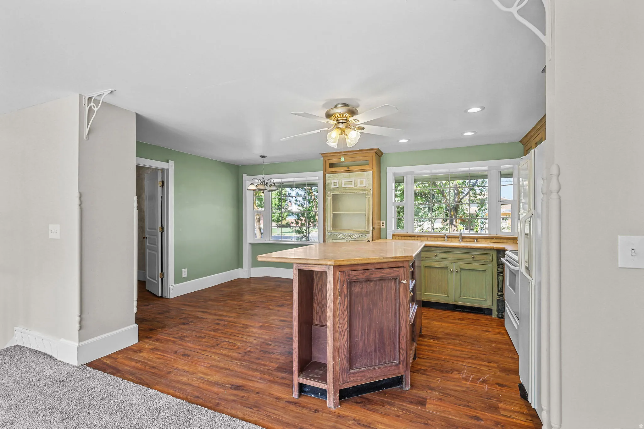 Wood-finish flooring, central kitchen island with countertop, recessed lighting, ceiling fan, and bay windows