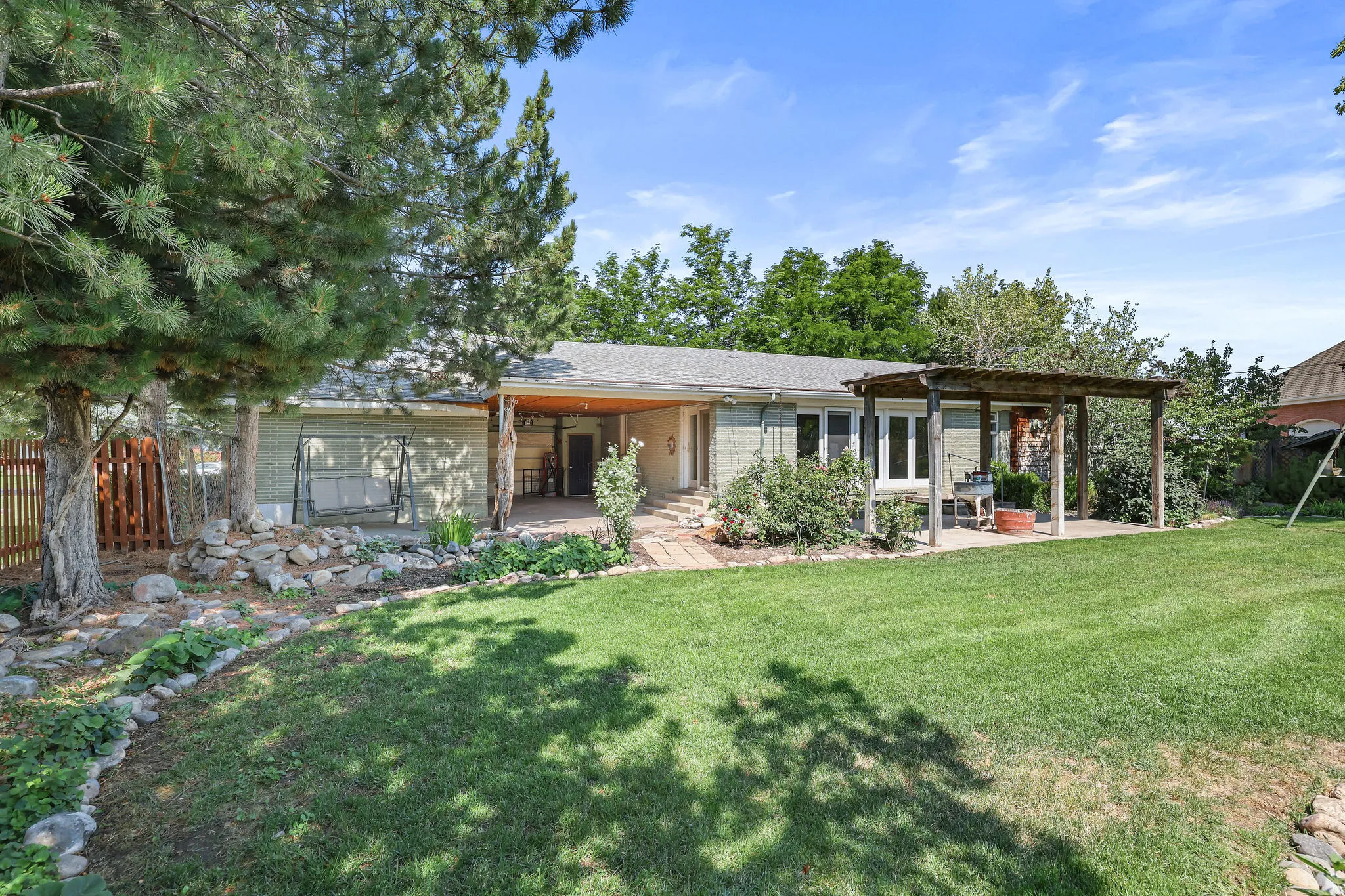 Expansive backyard with manicured lawn and mature tree coverage, anchored by a single-story residence featuring a covered patio, wood-beam pergola, and multi-panel sliding glass doors providing indoor-outdoor connectivity