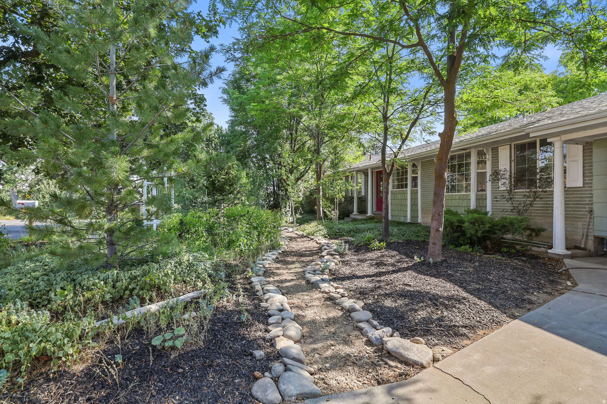 Exterior featuring green siding, white window shutters, mature trees, a stone-lined pathway, and a concrete driveway