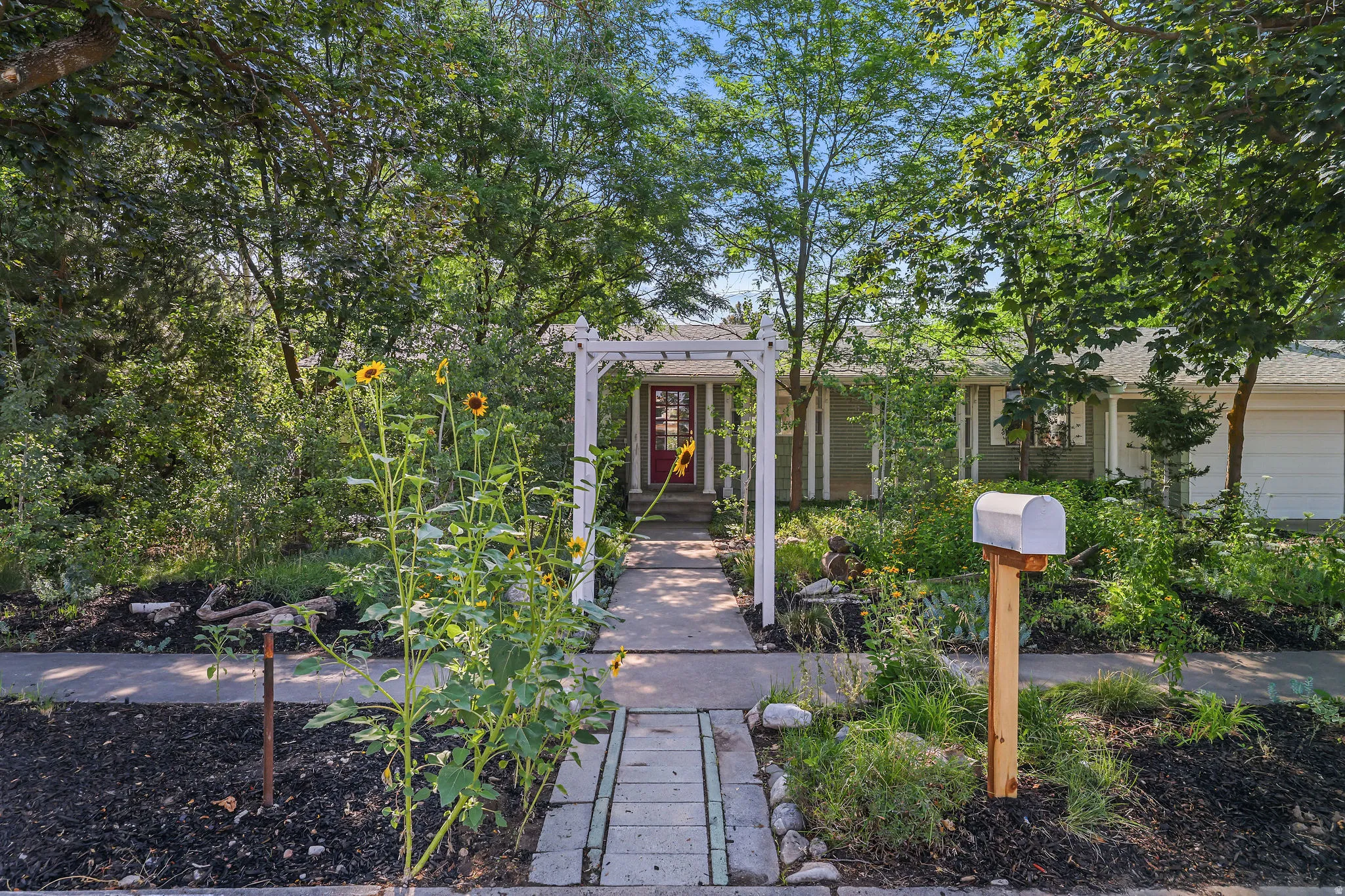 Front exterior featuring mature trees, a concrete pathway, a white arbor, a red entry door, and an attached garage